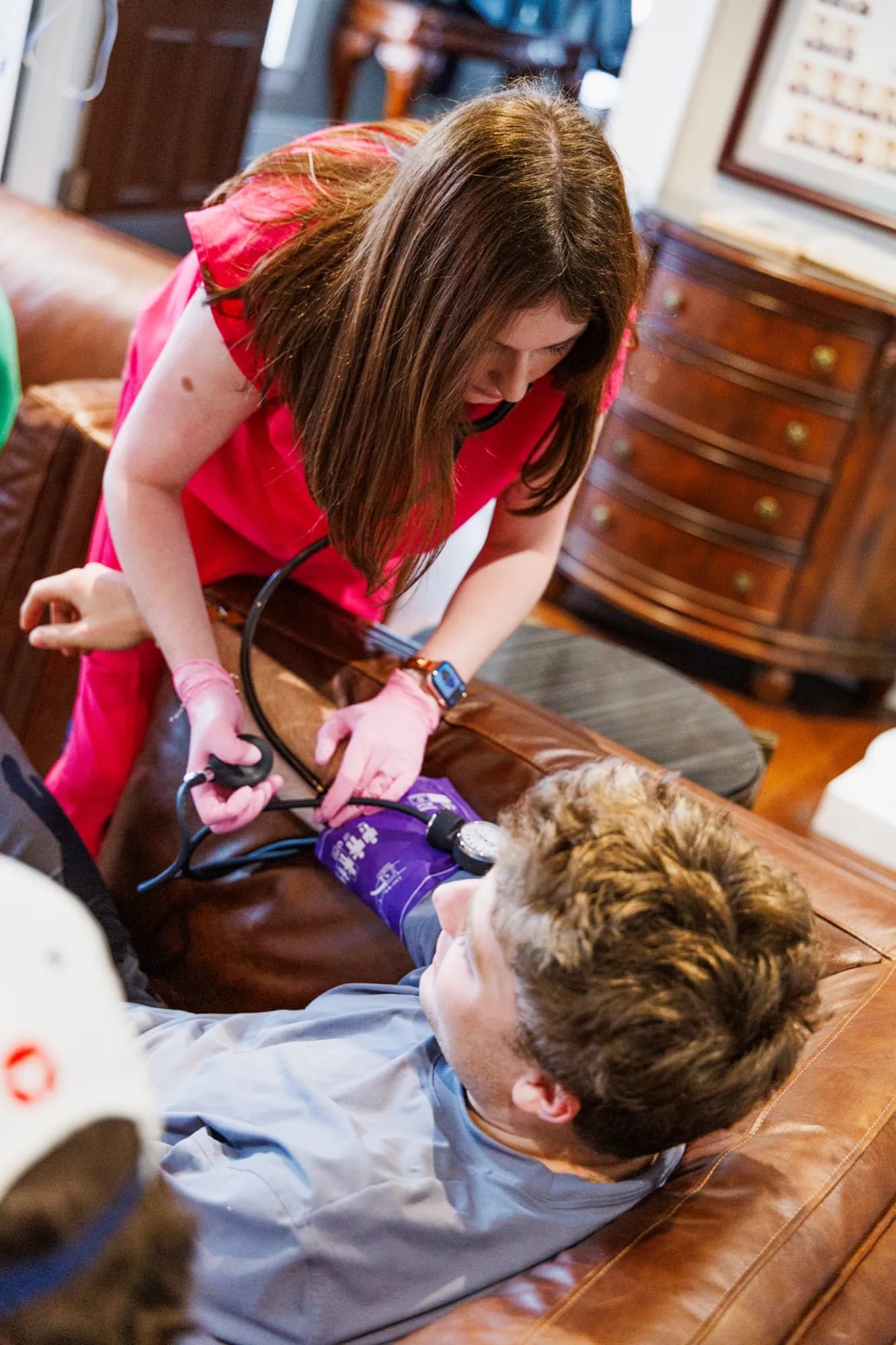 A ThrIVe registered nurse in pink scrubs starting an IV for a client on a leather couch